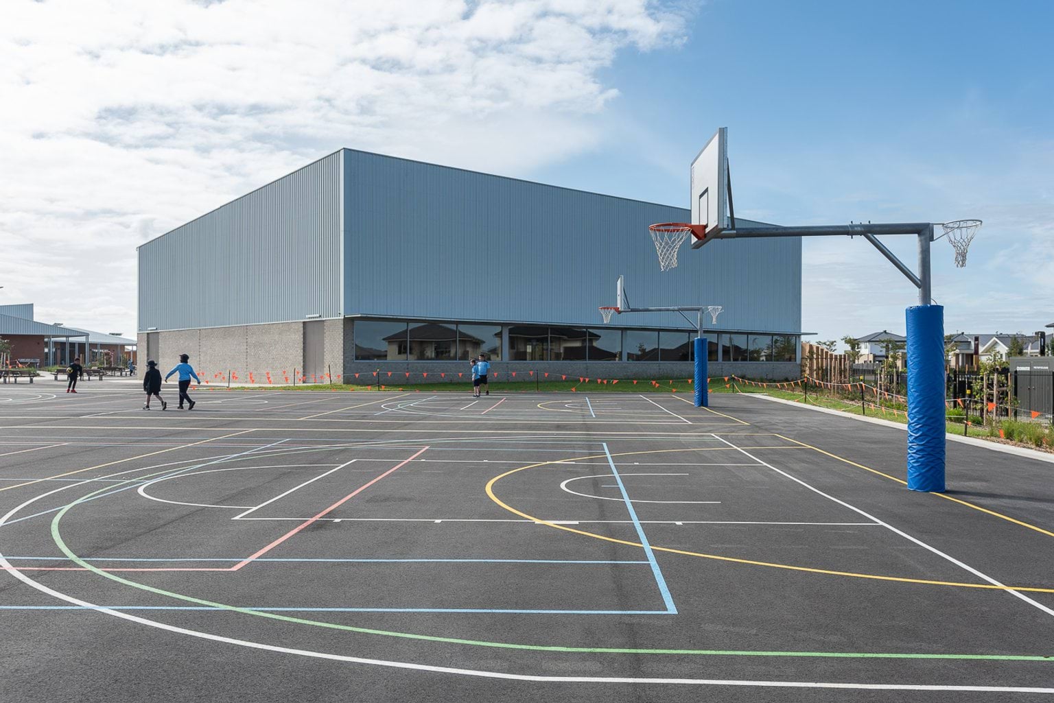 Outdoor space showing one quarter of a basketball court and a ring. In the background is a large gymnasium building.