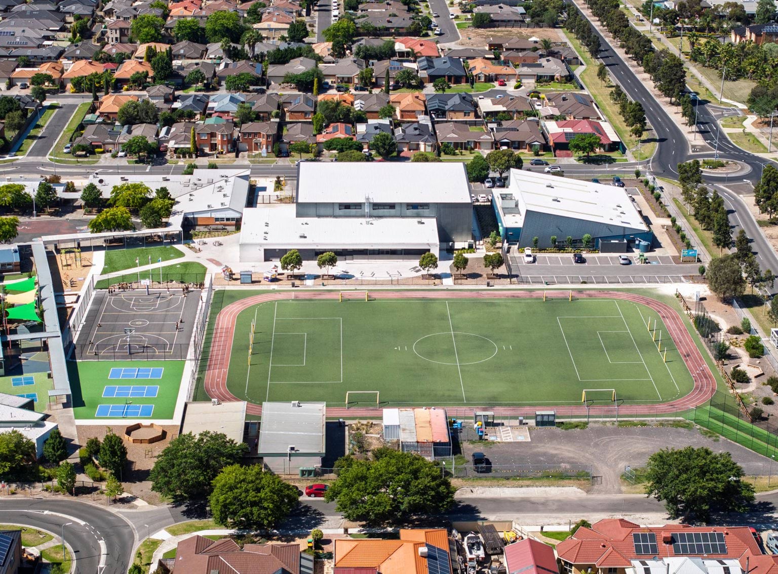 Birdseye view of a school, its athletics precinct and surrounding homes. There is a large oval with a soccer pitch which is surrounded by a red athletics track. There are basketball and tennis facilities to the left and the school building at the top of the image.