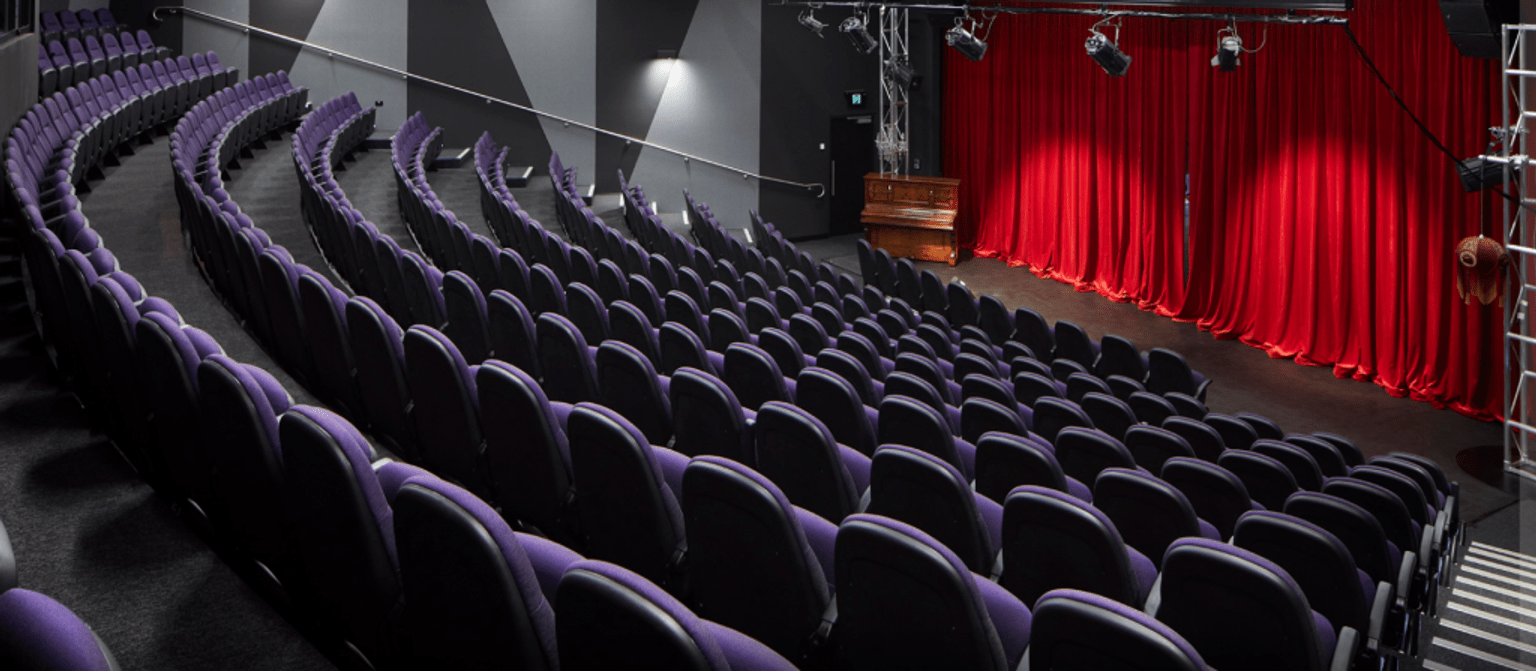 Image shows tiered purple seating in a theatre with a stage down at the front. The stage has a red curtain with two spotlights.