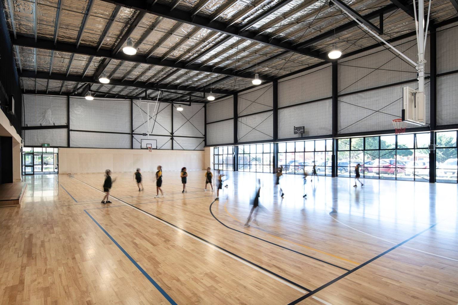 Single sized competition grade gymnasium with wooden floor. One side of the gym is flanked with glass windows making it a bright space. School aged children are blurred with movement.
