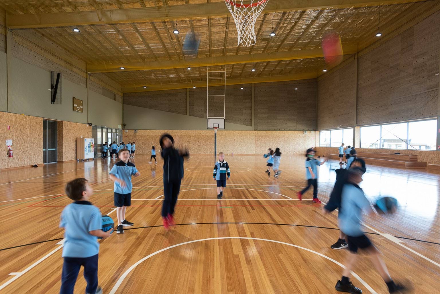 Single sized competition grade gymnasium with wooden floor. Primary school aged children are playing with basketballs. Students are blurred in movement, wearing light blue uniforms.